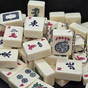 May include: A pile of off-white Mahjong tiles, some with keyrings attached. The tiles display red, black, and green symbols and numbers. One tile reads "JOKER" with a dragon design. The tiles are arranged in a random pattern.