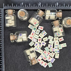 May include: Miniature glass bottles with cork stoppers, each containing small, off-white Mahjong tiles. Scattered around the bottles are additional tiles with colorful symbols and characters. The background is a dark surface with a ruler.