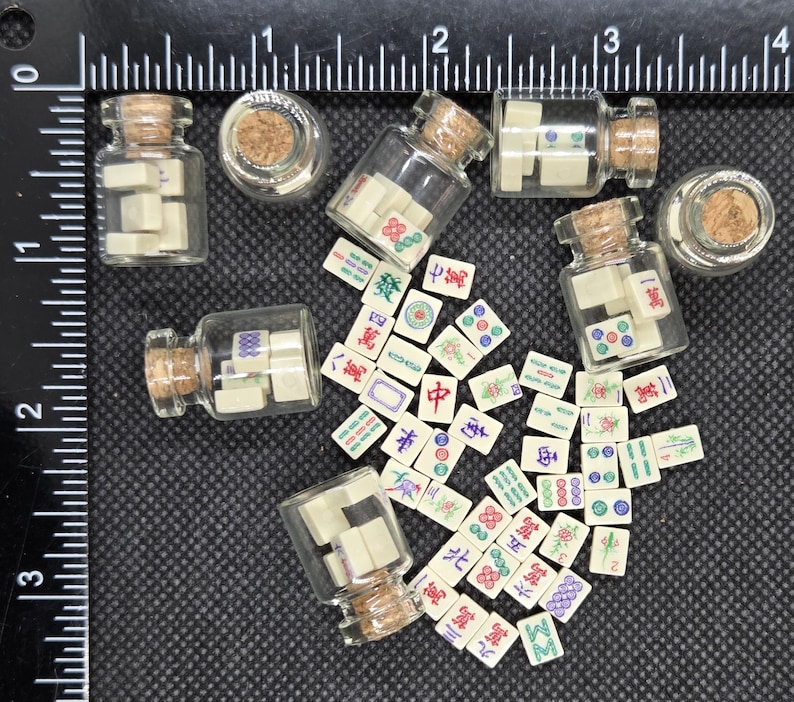 May include: Miniature glass bottles with cork stoppers, each containing small, off-white Mahjong tiles. Scattered around the bottles are additional tiles with colorful symbols and characters. The background is a dark surface with a ruler.