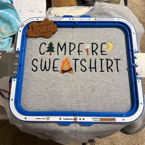 May include: A grey sweatshirt secured in a blue embroidery hoop featuring the embroidered text CAMPFIRE SWEATSHIRT in black. The design includes a green pine tree, a marshmallow on a stick, a yellow crescent moon, and a glowing orange campfire.