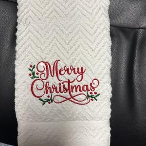 May include: White hand towel with a textured chevron pattern. The words "Merry Christmas" are embroidered in red script with green holly accents. A festive holiday themed towel.
