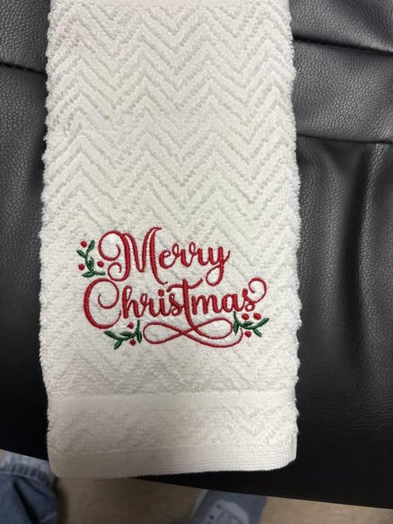 May include: White hand towel with a textured chevron pattern. The words "Merry Christmas" are embroidered in red script with green holly accents. A festive holiday themed towel.