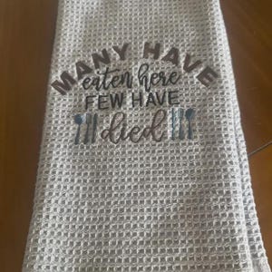 May include: A light gray waffle-weave kitchen towel with the embroidered text "MANY HAVE eaten here FEW HAVE died". The text is in a dark gray color, with a fork and spoon design.