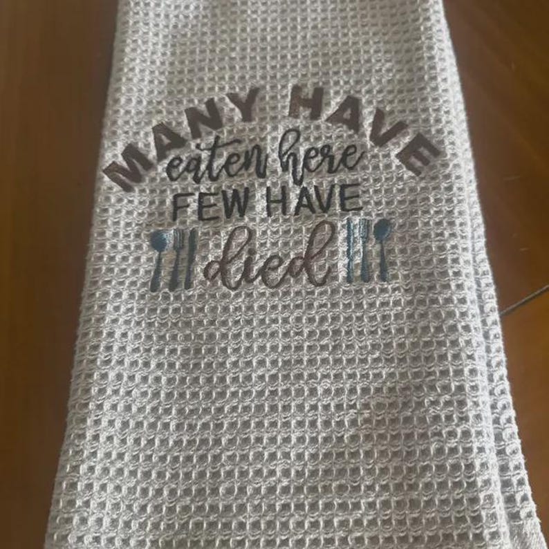 May include: A light gray waffle-weave kitchen towel with the embroidered text "MANY HAVE eaten here FEW HAVE died". The text is in a dark gray color, with a fork and spoon design.