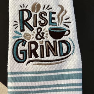 May include: White kitchen towel with the embroidered phrase "Rise & Grind" in teal and brown. The towel features coffee bean and cup illustrations. Teal and white striped border at the bottom.