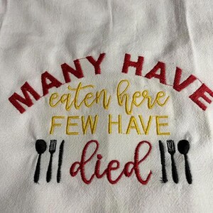 May include: White kitchen towel with embroidered text in red and yellow. The text reads "MANY HAVE eaten here FEW HAVE died" with black utensil graphics. A humorous and decorative kitchen accessory.