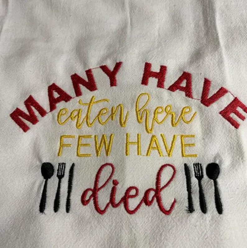 May include: White kitchen towel with embroidered text in red and yellow. The text reads "MANY HAVE eaten here FEW HAVE died" with black utensil graphics. A humorous and decorative kitchen accessory.