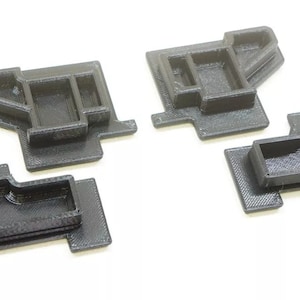 May include: Four black, 3D-printed plastic components. Each piece has a rectangular base with a raised, box-like structure on top. The components are arranged on a white surface, showcasing their geometric design and textured surfaces.