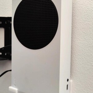 May include: A white Xbox Series S game console mounted vertically on a wall using a white plastic bracket system. The console features a prominent circular black ventilation grille on the front, a small Xbox logo, and a front-facing port and button.