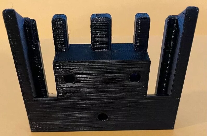May include: Black plastic 3D printed object with three vertical slots and four holes. The object has a rectangular shape.