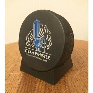 May include: A black hockey puck with a white logo that says "Steam Whistle" and "Canada's Premium Pilsner". The logo also features a blue and white illustration of a steam whistle.