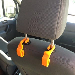 May include: Two orange plastic hooks hang from the headrests of a car seat. The hooks are designed to hang bags or other items from the back of the seat.