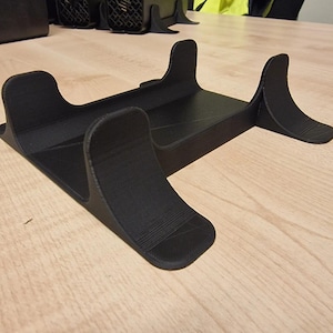 May include: A black, 3D-printed stand with a rectangular base and curved supports. The stand is designed to hold an electronic device. The supports are curved and angled, providing stability. The stand is made of a matte black material.