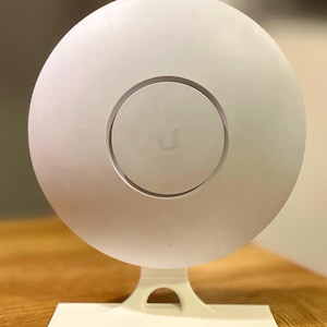 May include: A white circular wireless access point mounted on a matching white stand. The device features a smooth, matte finish with a central circular emblem containing the Ubiquiti logo. The stand has a wide, flat base and a curved support arm.