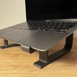 May include: A black laptop stand with a laptop on top of it. The stand is made of metal and has a simple design. The laptop is a silver MacBook Air.