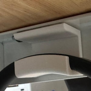 May include: Close-up of a white bracket attached to a metal frame, with a black circular object partially visible. The bracket appears to be supporting the object. A wooden surface is visible above.