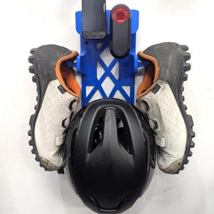 May include: A blue plastic wall-mounted storage rack holding a black bicycle helmet, a pair of white and orange cycling shoes, a black Wahoo cycling computer, and a black rear bicycle light. The rack features a geometric lattice design.