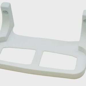 May include: A white plastic support bracket with two rectangular openings and two vertical posts with holes. The bracket has a curved design and is likely used for mounting or supporting an object. The overall shape is symmetrical.