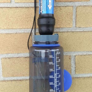 May include: A blue water filtration system with a Sawyer filter attached to a clear Nalgene water bottle. The filter has a blue label with the word "FLOW" and an arrow pointing down. The bottle has a blue lid and a blue measurement scale on the side. The system is hanging from a brick wall.