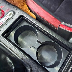 May include: A black plastic dual cup holder insert situated in a vehicle center console. The console features a red and black leather seat edge, a red seatbelt release button, and a control panel with text including S# Sport Sharp and C.DIFF.