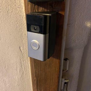 May include: A silver and black doorbell camera mounted on a wooden door frame. The camera has a round button and a lens.