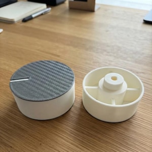 May include: Two circular components, one featuring a dark gray textured top with a thin slot and white sides, and the other an off-white open cylindrical base with a central raised hub and internal support ribs, both showing 3D printed construction.