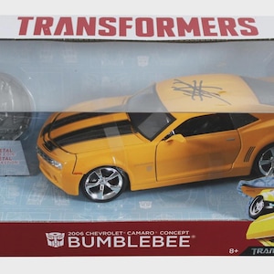 May include: A yellow 2006 Chevrolet Camaro Bumblebee concept car toy with black racing stripes, displayed in a clear plastic box. The box features the Transformers logo and text. The toy is made of 100% metal.