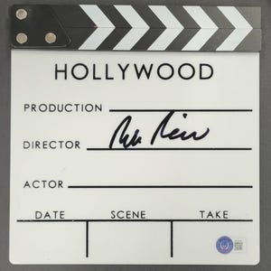 May include: A white and black film clapperboard with the word "HOLLYWOOD" printed at the top. The clapperboard has lines for production, director, actor, date, scene, and take. A signature is present on the director's line.