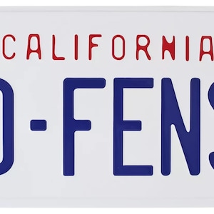 Puede incluir: Una placa de matrícula blanca de California con letras azules que dicen "D-FENS". El nombre del estado "CALIFORNIA" está en letras rojas. La placa está firmada con tinta azul por "The Dude".