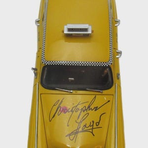May include: A yellow toy taxi cab viewed from above. The cab features a black and white chequered pattern above the windscreen and the name "Christopher Hayd" in black script on the bonnet. A roof sign reads "On Hand Sale".