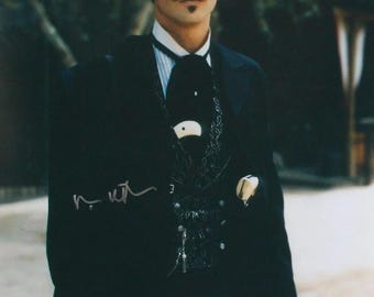 May include: A person wearing a black wide-brimmed hat, a black suit jacket, a black patterned vest, and a light blue and white striped shirt. A white-handled pistol is tucked into the vest, and a signature is visible on the jacket.