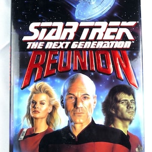 May include: A book cover for the Star Trek: The Next Generation novel "Reunion" by Michael Jan Friedman. The cover features a starship against a dark blue background with stars. The title is in red and white lettering. The cover also features three characters from the show: Captain Jean-Luc Picard, Deanna Troi, and Worf.