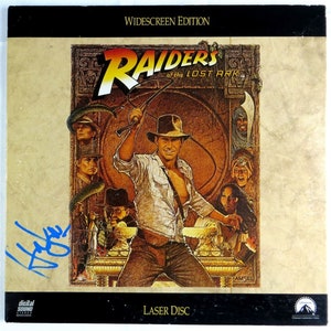 Könnte beinhalten: Ein Vintage-Laserdisc-Etui für den Film Jäger des verlorenen Schatzes. Das Cover zeigt eine farbenfrohe Illustration von Indiana Jones, der eine Peitsche hält, umgeben von anderen Charakteren aus dem Film. Das Etui ist mit "Widescreen Edition" und "Laser Disc" beschriftet.