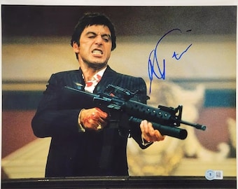 May include: A black and white photo of a man holding a gun. He is wearing a black suit and a white shirt. The man is looking at the camera with a serious expression. The photo is signed in blue ink.