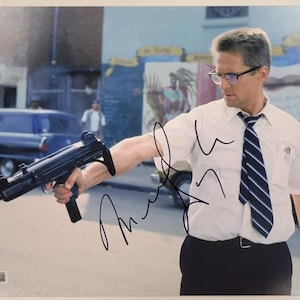 May include: A signed photograph featuring a man in a white shirt and tie holding a black submachine gun. The man is wearing glasses and is in an outdoor setting. The photograph has a signature in black ink.