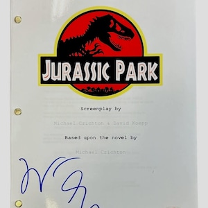 Puede incluir: Un guion blanco con el logo de "Jurassic Park" en rojo, negro y amarillo. El guion está firmado con tinta azul. El texto dice "Guion de Michael Crichton & David Koepp". Una pegatina de autenticación PSA está en la esquina inferior derecha.
