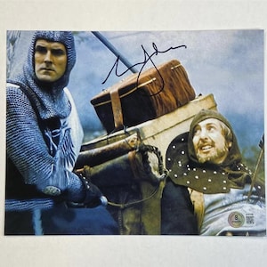 May include: A signed photograph featuring two men in medieval attire. One man is in chainmail armor, and the other wears a hooded cloak. The image has a vintage aesthetic, with a signature in the upper right corner.