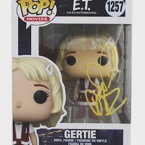 May include: Funko Pop! Movies figure of Gertie from E.T. The Extra-Terrestrial. The figure is in a box with the Funko Pop! and E.T. logos. The figure is wearing a red and white dress and has blonde hair. The box has a yellow signature.