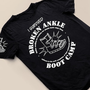 May include: Black t-shirt with a white graphic of a broken ankle in a boot. The graphic says "I Survived Broken Ankle Boot Camp".