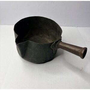 May include: Antique copper saucepan with a long, cylindrical handle. The pan has a dark patina, showing signs of age and use. The handle is a contrasting brown color, adding to the vintage aesthetic. The saucepan is designed with a pouring spout.