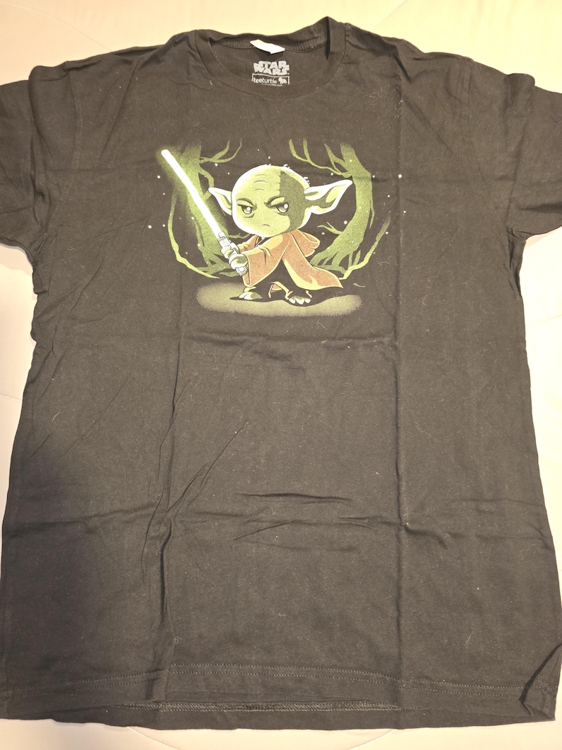 Puede incluir: Camiseta negra de Star Wars con un dise&ntilde;o de Yoda de dibujos animados sosteniendo un sable de luz verde. El dise&ntilde;o incluye un fondo de bosque. La camiseta tiene cuello redondo y mangas cortas.