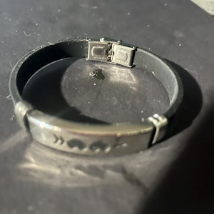 May include: A black leather bracelet with a silver clasp and a silver plate with three heart symbols. The bracelet is approximately 8 inches (20 cm) in circumference.