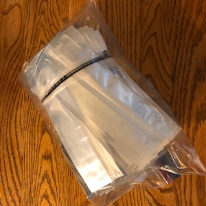 May include: A clear plastic bag filled with a large quantity of small, clear, rectangular pouches. The pouches are made of a shiny, silver-colored material.