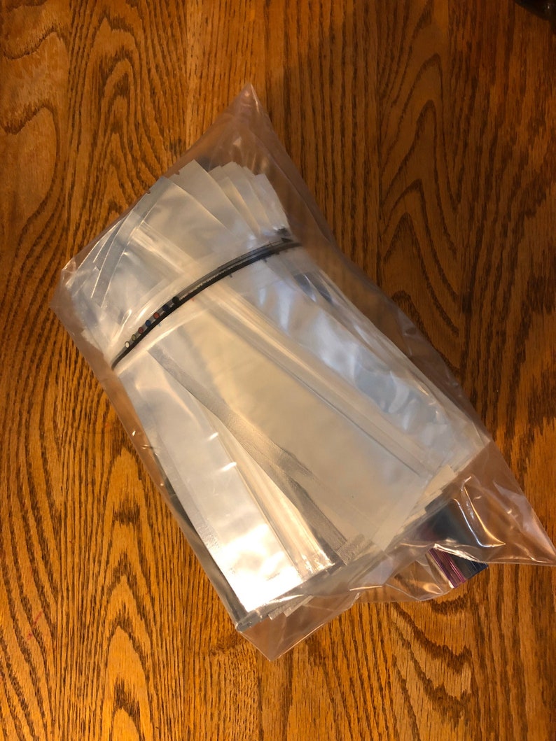May include: A clear plastic bag filled with a large quantity of small, clear, rectangular pouches. The pouches are made of a shiny, silver-colored material.