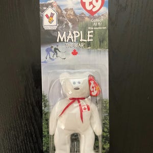 May include: A white plush bear toy named "Maple" with a red ribbon and a Canadian flag emblem. The packaging features a scenic background with mountains, trees, and the text "MAPLE THE BEAR". The toy is in a clear plastic case.