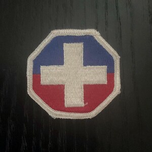 May include: A white cross patch with a blue top and red bottom, outlined in white. The patch has a white border and an octagonal shape. The patch is likely an emblem or insignia.