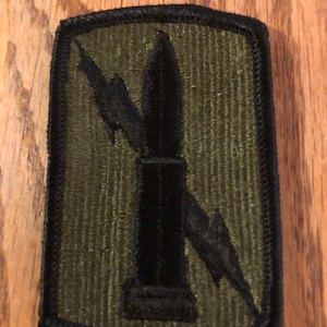 May include: A green military patch with a black silhouette of a rocket and lightning bolts.