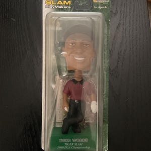 Puede incluir: Una figura de bobblehead de Tiger Woods en un embalaje de plástico transparente. La figura lleva una camisa granate, pantalones negros y una gorra negra. El embalaje dice "TIGER SLAM" y "2000 PGA Championship."