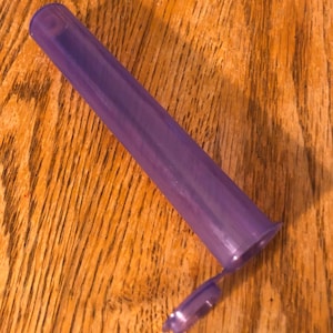 May include: A clear, purple plastic tube with a lid. The tube is about 10 inches long and 1 inch in diameter.