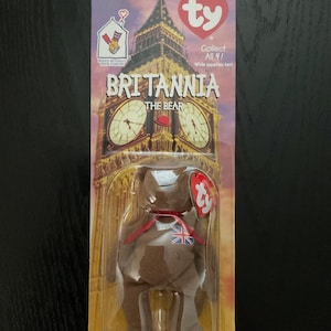 Peut inclure: Un ours en peluche Britannia de la collection Beanie Baby dans son emballage d'origine. L'emballage présente une image de Big Ben et le texte "BRITANNIA THE BEAR". L'ours porte un ruban rouge et un drapeau britannique sur la poitrine.